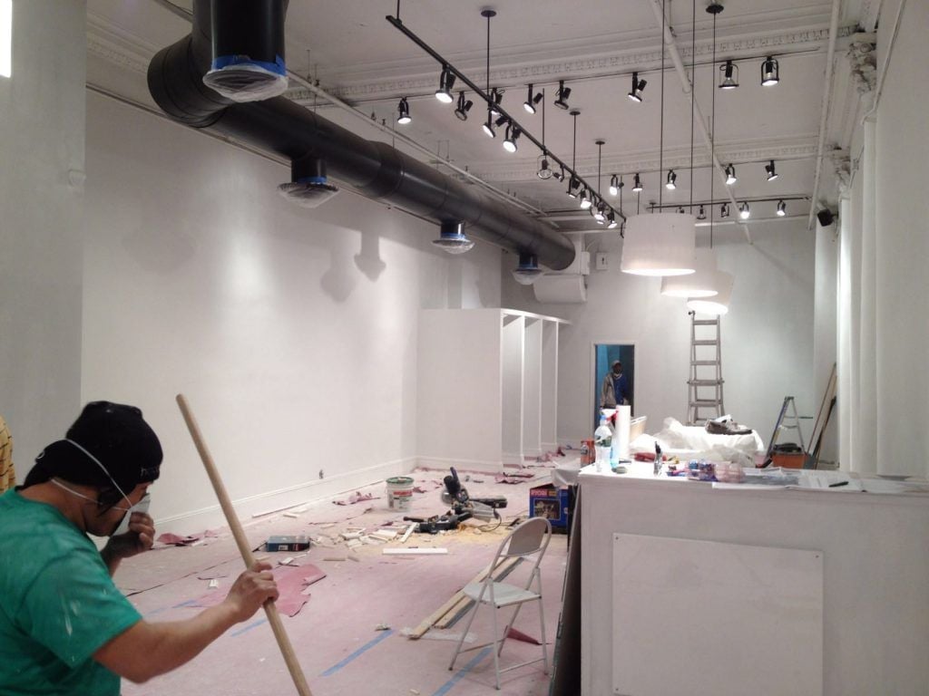 Construction worker in green shirt painting walls inside an empty industrial warehouse space with track lighting and white walls